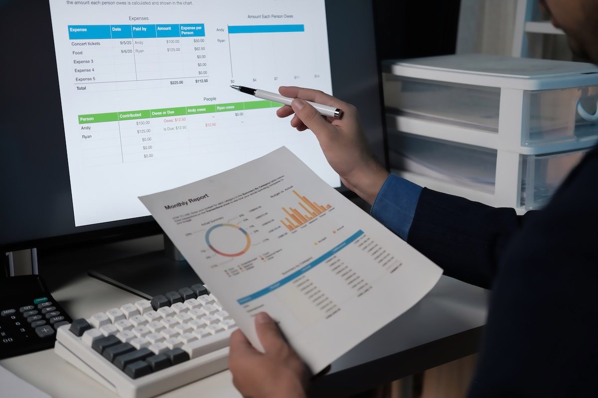 A person is looking at a computer screen with a graph on it and holding a piece of paper. The person is pointing at the graph and seems to be explaining something to someone else